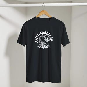 May include: Black t-shirt with a white graphic of a clenched fist and the words "ANTI-NOWHERE LEAGUE" in a circular design. The shirt is displayed on a wooden hanger.