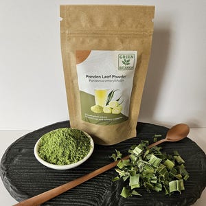Könnte beinhalten: Ein brauner Kraftpapierbeutel mit der Aufschrift Green Botanica Pandan Leaf Powder Pandanus amaryllifolius. Daneben stehen eine kleine weiße Schale mit leuchtend grünem Pulver, ein Haufen getrockneter Pandanblätter und ein langer Holzlöffel.