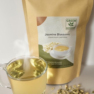 Peut inclure: Une tasse en verre remplie de th&eacute; dor&eacute;, de la vapeur s'en d&eacute;gage, &agrave; c&ocirc;t&eacute; d'un sachet en papier kraft de Fleurs de Jasmin. Le sac porte une &eacute;tiquette avec le texte "Green Botanica" et "Fleurs de Jasmin". Des fleurs de jasmin s&eacute;ch&eacute;es sont &eacute;parpill&eacute;es &agrave; proximit&eacute;.