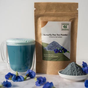 Puede incluir: Una bolsa de papel marrón etiquetada Green Botanica Butterfly Pea Tea Powder Clitoria ternatea, una taza de cristal llena de té azul vibrante con espuma y un cuenco blanco pequeño con un montón de polvo azul, rodeado de flores azules.