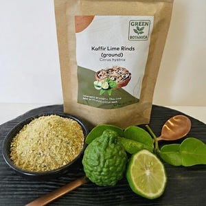 May include: A kraft paper bag of ground Kaffir lime rinds with the text "GREEN BOTANICA". A small bowl of ground lime rinds, a whole lime, a lime half, and a copper spoon are in the foreground. Green leaves and a whole lime are also visible.