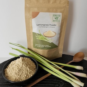 May include: A brown paper bag of lemongrass powder with a green and white label that reads "Lemongrass Powder". A black bowl of lemongrass powder, fresh lemongrass stalks, and a wooden spoon are in the foreground. The product is from Green Botanica.