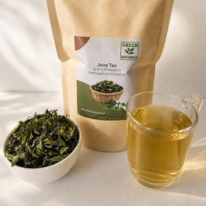 Könnte beinhalten: Ein brauner Papierbeutel mit der Aufschrift Green Botanica Java Tea Cat's Whiskers Orthosiphon aristatus, eine weiße Schale mit getrockneten grünen Kräuterblättern und eine klare Glastasse mit einem hellgoldenen Tee auf einer Fläche.