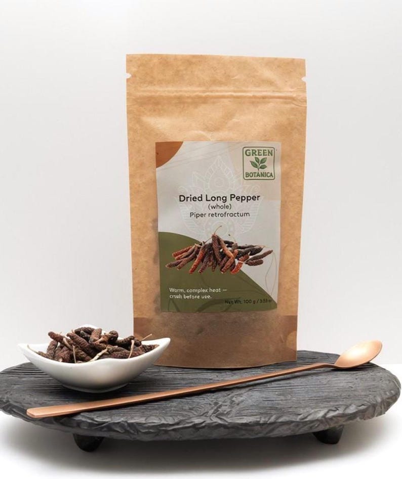 May include: A brown paper bag of dried long pepper with the text "Dried Long Pepper (whole) Piper retrofractum". A small white bowl of dried long pepper and a copper spoon are on a black, round, wooden tray.