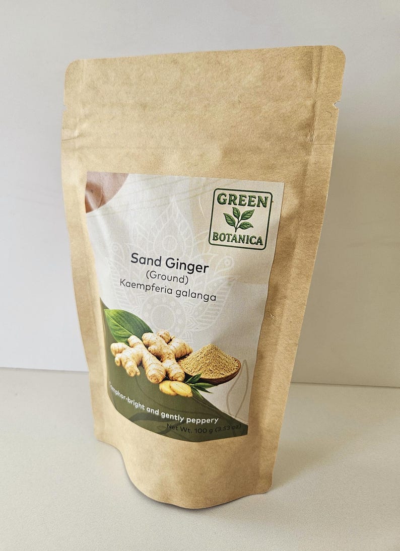 Op de afbeelding: Een bruine papieren zak met gemalen zandgember, gelabeld Green Botanica, Kaempferia galanga. Het etiket toont een afbeelding van verse gemberwortel en een klein kommetje poeder, met de tekst Net Wt. 100 g onderaan afgedrukt.