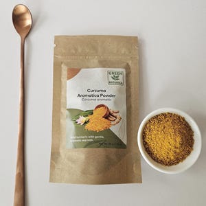 May include: A brown paper bag of "Curcuma Aromatica Powder" and a small white bowl filled with yellow powder. A long, copper-coloured spoon rests to the left. The bag has text that reads "Wild turmeric with gentle, aromatic warmth."