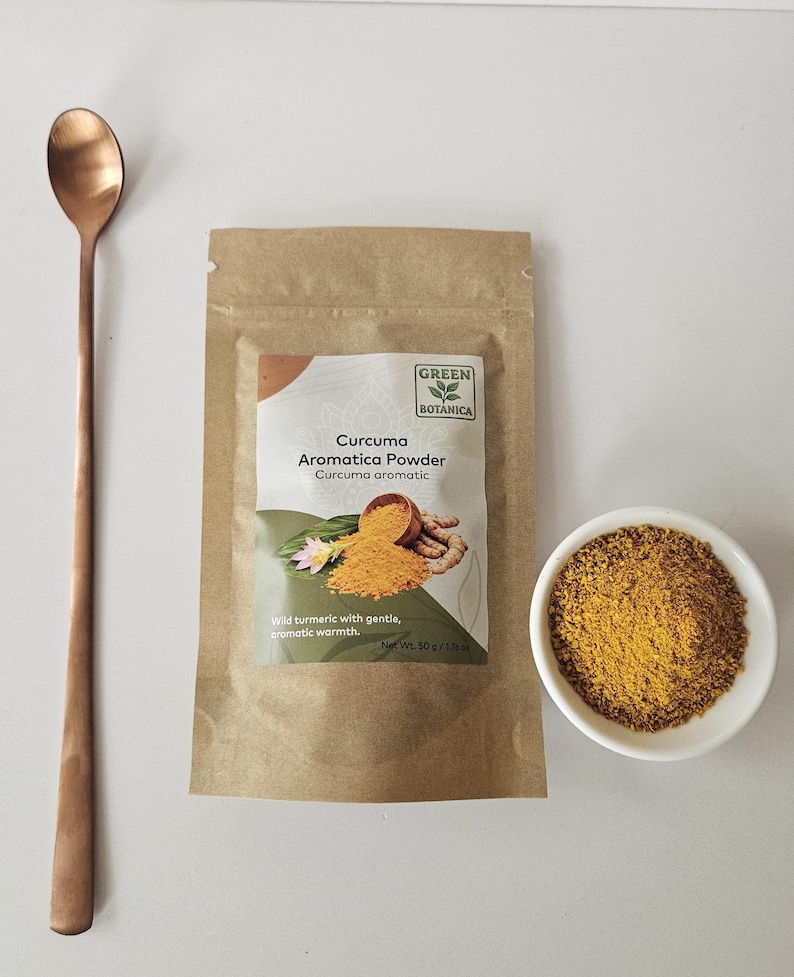 May include: A brown paper bag of "Curcuma Aromatica Powder" and a small white bowl filled with yellow powder. A long, copper-coloured spoon rests to the left. The bag has text that reads "Wild turmeric with gentle, aromatic warmth."