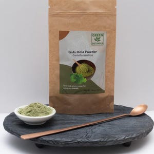 May include: A brown paper bag of Gotu Kola Powder with a label featuring a bowl of green powder and the words "Green Botanica". A small white bowl and a copper spoon sit on a dark grey, round, raised tray. The bowl contains green powder.