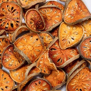 May include: Close-up of dried bael fruit slices. The slices are a mix of orange and brown hues, with a textured appearance and visible seed patterns. The fruit is arranged in a pile, showcasing the natural variations in size and shape.