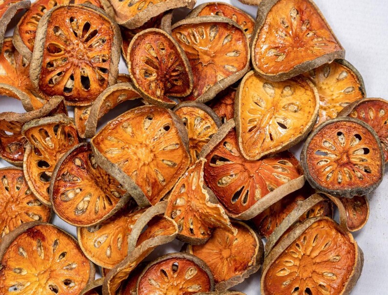 May include: Close-up of dried bael fruit slices. The slices are a mix of orange and brown hues, with a textured appearance and visible seed patterns. The fruit is arranged in a pile, showcasing the natural variations in size and shape.