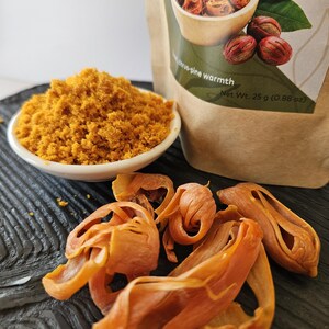 K&ouml;nnte beinhalten: Eine kleine wei&szlig;e Schale mit goldbraunem gemahlenem Muskatbl&uuml;ten-Gew&uuml;rz, begleitet von mehreren ganzen getrockneten Muskatbl&uuml;ten und einem braunen Papierbeutel mit der Aufschrift citrus-pine warmth und Net Wt. 25 g (0.88 oz).