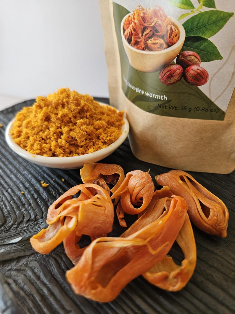 K&ouml;nnte beinhalten: Eine kleine wei&szlig;e Schale mit goldbraunem gemahlenem Muskatbl&uuml;ten-Gew&uuml;rz, begleitet von mehreren ganzen getrockneten Muskatbl&uuml;ten und einem braunen Papierbeutel mit der Aufschrift citrus-pine warmth und Net Wt. 25 g (0.88 oz).