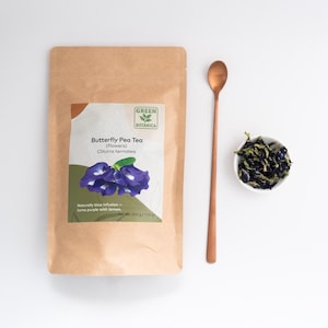 Pode incluir: Uma embalagem de papel castanho de ch&aacute; de flores de feij&atilde;o-borboleta Green Botanica com um r&oacute;tulo que mostra flores azuis e o texto Net Wt. 200 g / 7.05 oz. Ao lado da embalagem h&aacute; uma colher de metal cor de cobre e uma ta&ccedil;a branca com flores secas.