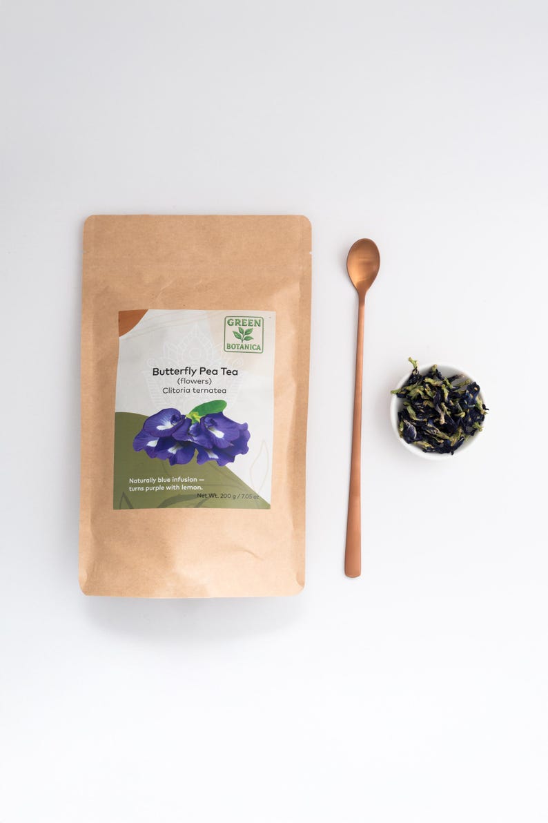 Pode incluir: Uma embalagem de papel castanho de ch&aacute; de flores de feij&atilde;o-borboleta Green Botanica com um r&oacute;tulo que mostra flores azuis e o texto Net Wt. 200 g / 7.05 oz. Ao lado da embalagem h&aacute; uma colher de metal cor de cobre e uma ta&ccedil;a branca com flores secas.