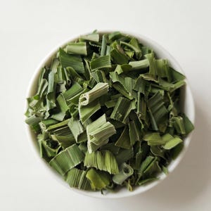 Może przedstawiać: Biała miska wypełniona posiekanymi zielonymi liśćmi pandanu. Liście mają żywy zielony kolor i wydają się świeżo ścięte. Miska jest wyśrodkowana na białym tle, tworząc czystą i prostą kompozycję.