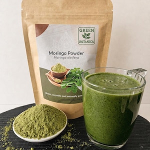Może przedstawiać: Brązowa papierowa torebka z etykietą Green Botanica Moringa Powder o wadze netto 100g, mała biała miseczka wypełniona zielonym proszkiem oraz przezroczysty szklany kubek zawierający gęste zielone smoothie.