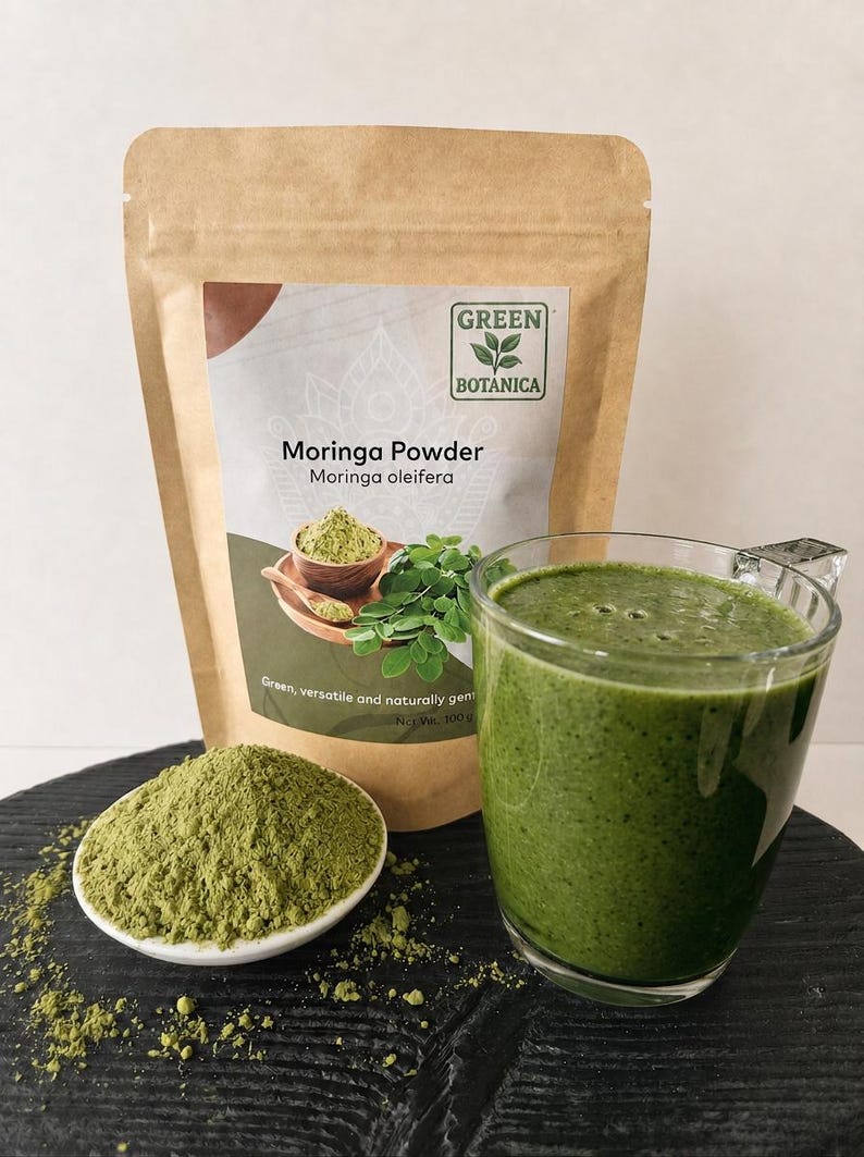 Może przedstawiać: Brązowa papierowa torebka z etykietą Green Botanica Moringa Powder o wadze netto 100g, mała biała miseczka wypełniona zielonym proszkiem oraz przezroczysty szklany kubek zawierający gęste zielone smoothie.