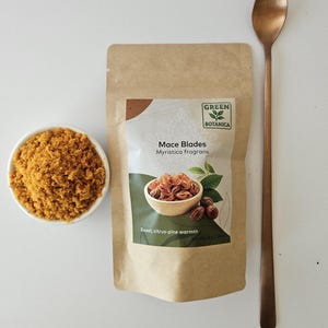 K&ouml;nnte beinhalten: Ein brauner Papierbeutel mit der Aufschrift Green Botanica Mace Blades Myristica fragrans und einem Nettogewicht von 25g, begleitet von einer kleinen wei&szlig;en Schale mit gemahlener Muskatbl&uuml;te und einem langen kupferfarbenen L&ouml;ffel.