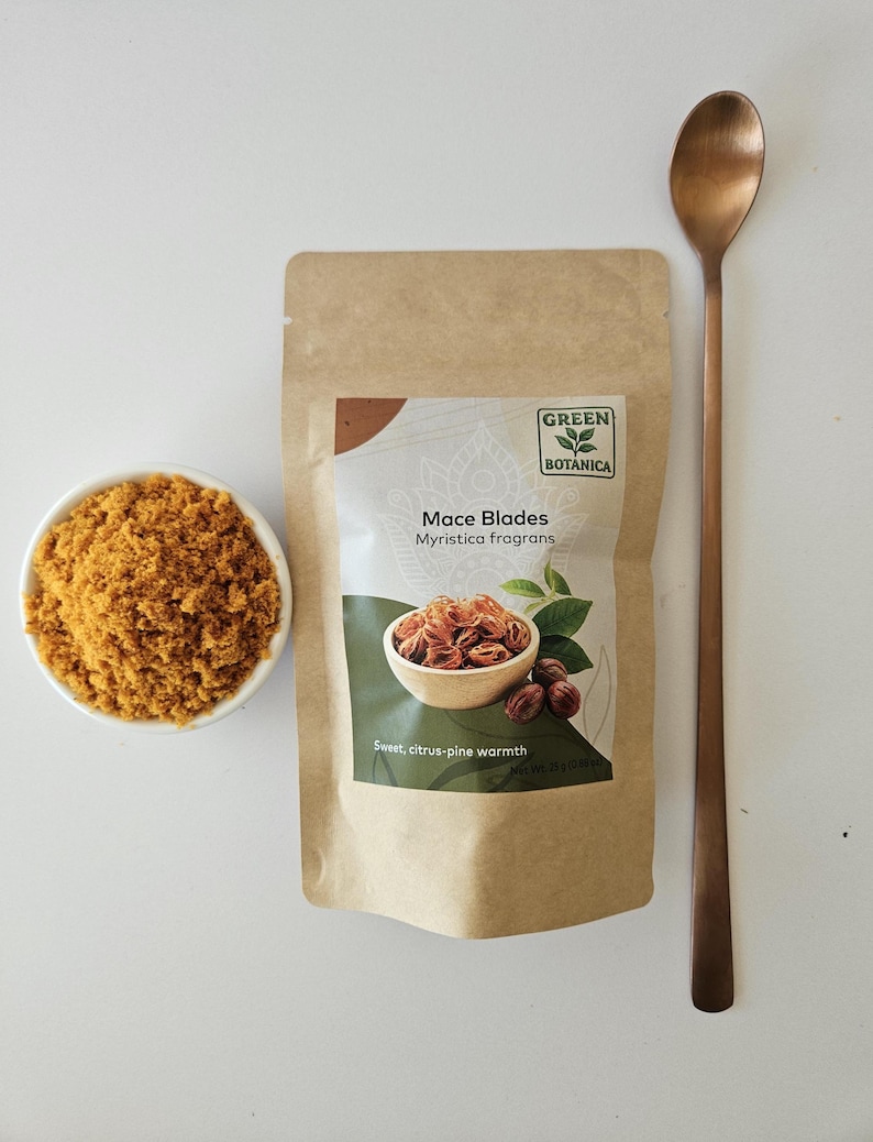 K&ouml;nnte beinhalten: Ein brauner Papierbeutel mit der Aufschrift Green Botanica Mace Blades Myristica fragrans und einem Nettogewicht von 25g, begleitet von einer kleinen wei&szlig;en Schale mit gemahlener Muskatbl&uuml;te und einem langen kupferfarbenen L&ouml;ffel.