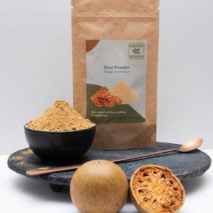 May include: A brown paper bag of Bael Powder with the text "Bael Powder Aegle marmelos" and a black bowl filled with powder. A whole bael fruit and a halved bael fruit are on a black tray with a copper spoon.