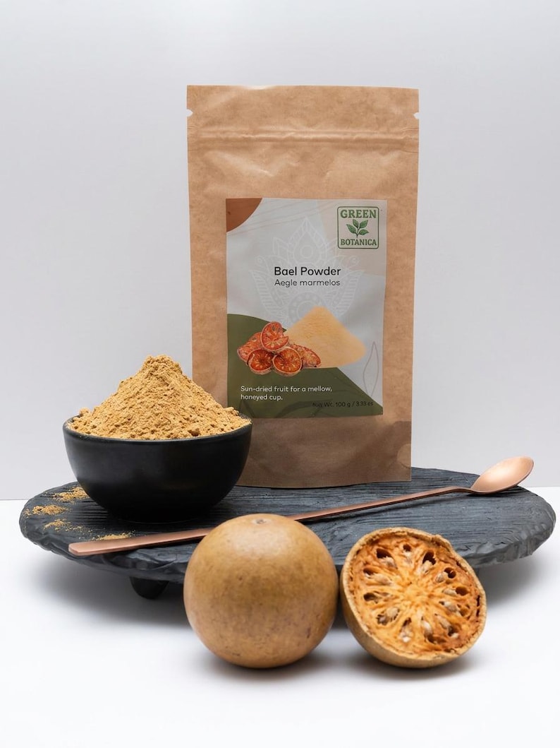 May include: A brown paper bag of Bael Powder with the text "Bael Powder Aegle marmelos" and a black bowl filled with powder. A whole bael fruit and a halved bael fruit are on a black tray with a copper spoon.