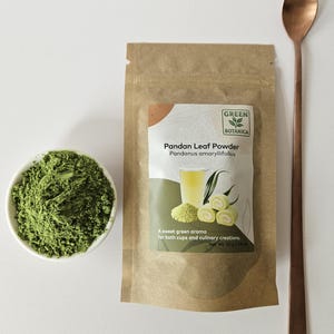 May include: A brown paper pouch of Green Botanica Pandan Leaf Powder with a label showing a glass of liquid and green rolls. Next to it is a small white bowl filled with green powder and a long-handled copper-colored metal spoon.