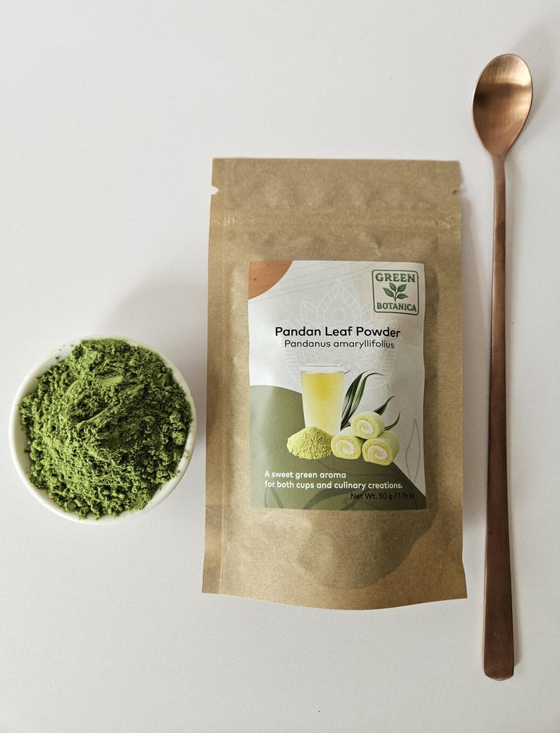 May include: A brown paper pouch of Green Botanica Pandan Leaf Powder with a label showing a glass of liquid and green rolls. Next to it is a small white bowl filled with green powder and a long-handled copper-colored metal spoon.
