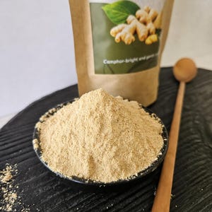 Op de afbeelding: Een kleine zwarte kom gevuld met lichtbruin zandgemberpoeder, geplaatst voor een bruine papieren zak met de tekst Sand Ginger (Ground) Kaempferia galanga en Camphor-bright and gentle, vergezeld van een houten lepel.