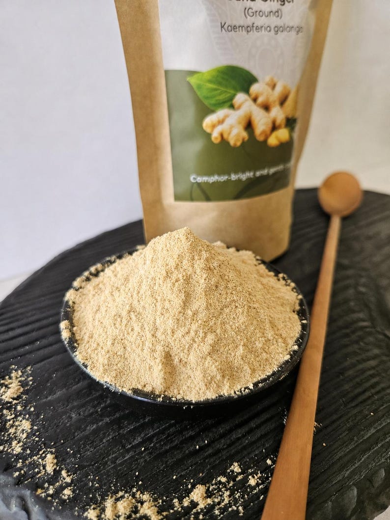 Op de afbeelding: Een kleine zwarte kom gevuld met lichtbruin zandgemberpoeder, geplaatst voor een bruine papieren zak met de tekst Sand Ginger (Ground) Kaempferia galanga en Camphor-bright and gentle, vergezeld van een houten lepel.