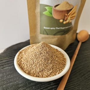 May include: A white bowl of ground fingerroot sits on a dark surface, with a wooden spoon beside it. A bag of fingerroot is in the background, with text that reads "Fingerroot (ground) Boesenbergia rotunda" and "Sweet-spicy Thai fingerroot".