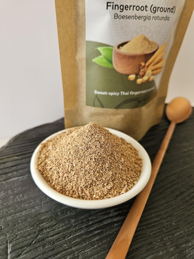 May include: A white bowl of ground fingerroot sits on a dark surface, with a wooden spoon beside it. A bag of fingerroot is in the background, with text that reads "Fingerroot (ground) Boesenbergia rotunda" and "Sweet-spicy Thai fingerroot".
