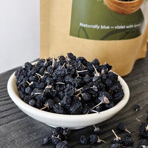 K&ouml;nnte beinhalten: Eine wei&szlig;e Keramikschale gef&uuml;llt mit getrockneten dunkelblauen Beeren, einige mit kleinen Stielen. Im Hintergrund befindet sich ein beigefarbener Papierbeutel mit einem gr&uuml;nen Etikett, das den Text Naturally blue - violet with lemon tr&auml;gt.