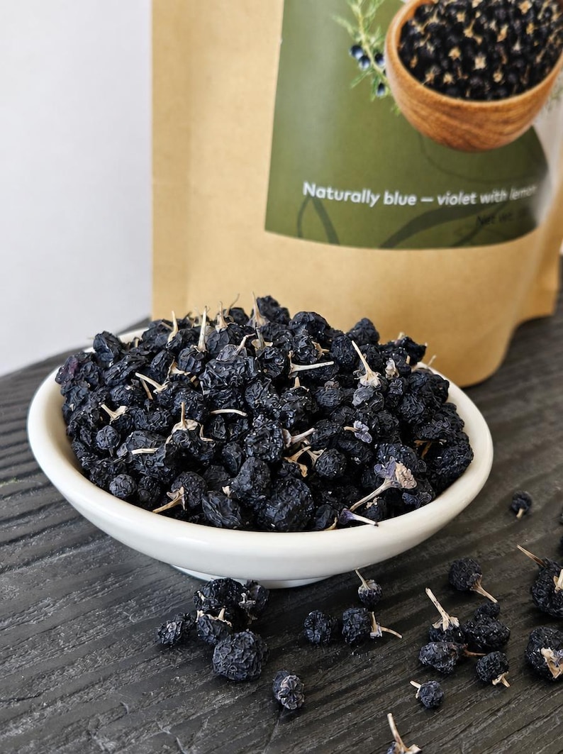 K&ouml;nnte beinhalten: Eine wei&szlig;e Keramikschale gef&uuml;llt mit getrockneten dunkelblauen Beeren, einige mit kleinen Stielen. Im Hintergrund befindet sich ein beigefarbener Papierbeutel mit einem gr&uuml;nen Etikett, das den Text Naturally blue - violet with lemon tr&auml;gt.