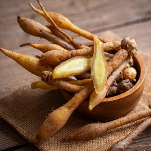May include: A wooden bowl filled with fresh fingerroot, also known as krachai. The fingerroot has a light brown skin and a yellow interior. The bowl sits on a burlap cloth, with a wooden background.