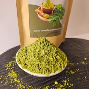 Może przedstawiać: Kopczyk jaskrawozielonego proszku Moringa znajduje się w małej białej miseczce. Za nią znajduje się beżowa torebka z etykietą zawierającą tekst Moringa Powder, Moringa oleifera oraz Green, versatile and naturally produced wraz z grafiką proszku.