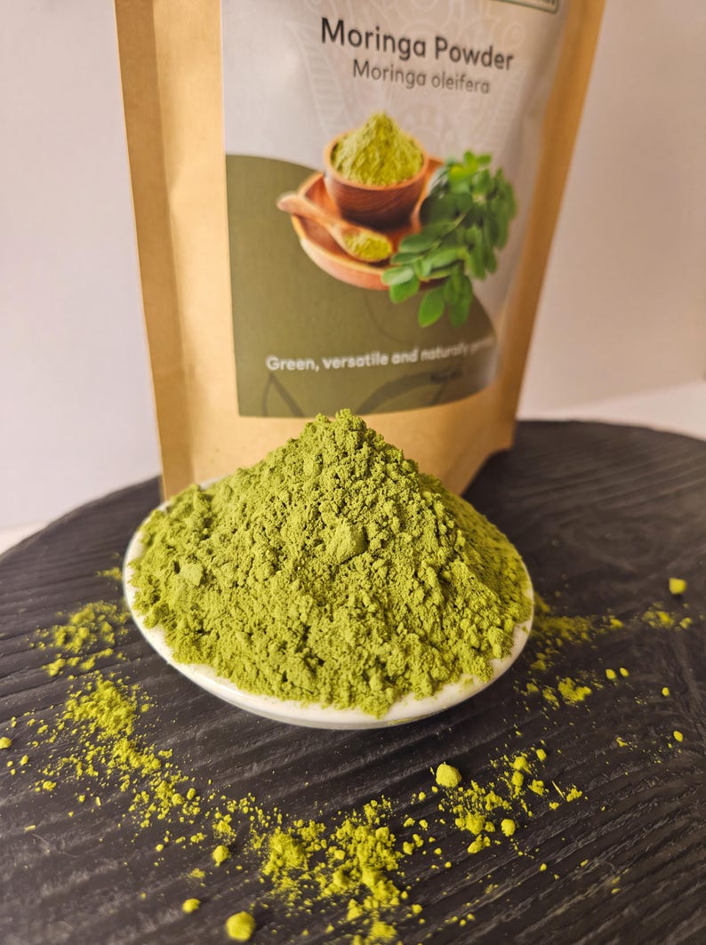 Może przedstawiać: Kopczyk jaskrawozielonego proszku Moringa znajduje się w małej białej miseczce. Za nią znajduje się beżowa torebka z etykietą zawierającą tekst Moringa Powder, Moringa oleifera oraz Green, versatile and naturally produced wraz z grafiką proszku.