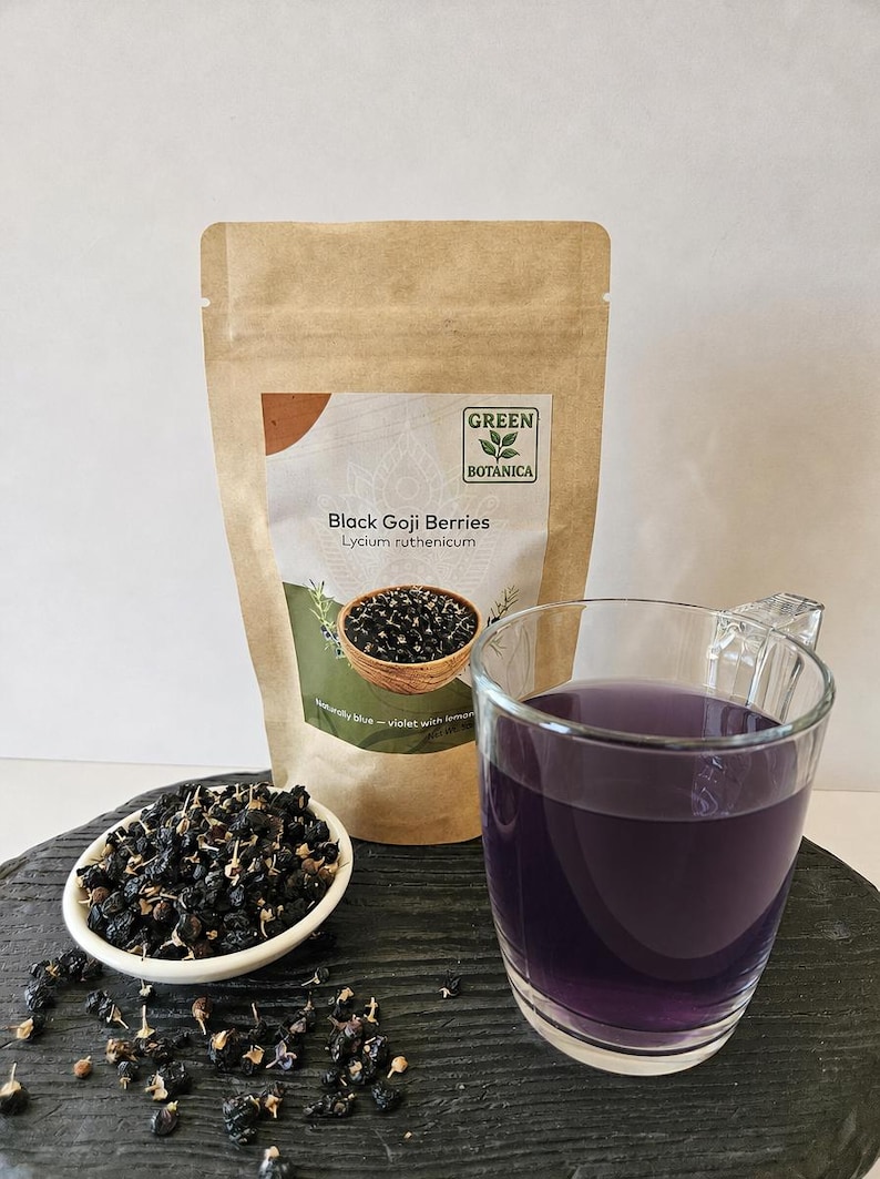 K&ouml;nnte beinhalten: Ein brauner Papierbeutel mit der Aufschrift Green Botanica Black Goji Berries Lycium ruthenicum, eine kleine wei&szlig;e Schale mit getrockneten schwarzen Goji-Beeren und eine klare Glastasse mit einem tiefvioletten Aufguss auf dunkler Oberfl&auml;che.