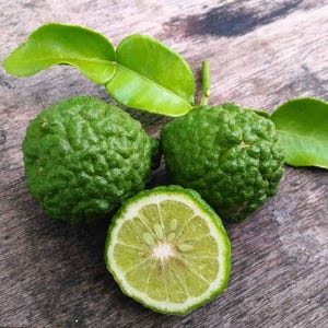 May include: A close-up shot of fresh green kaffir limes and leaves on a wooden surface. One lime is sliced in half, showing the inner segments. The textured skin of the limes and the vibrant green leaves create a natural, organic aesthetic.