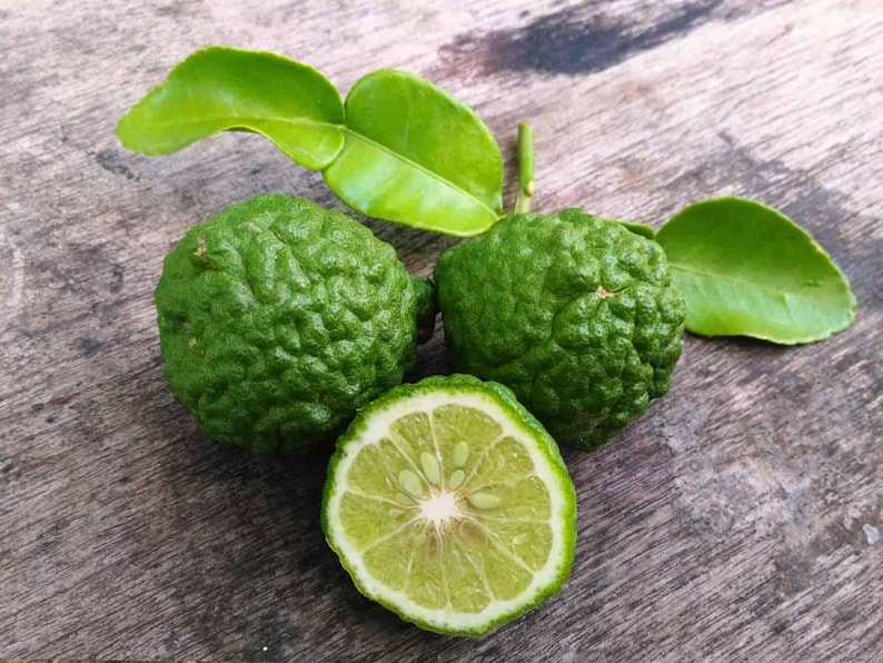 May include: A close-up shot of fresh green kaffir limes and leaves on a wooden surface. One lime is sliced in half, showing the inner segments. The textured skin of the limes and the vibrant green leaves create a natural, organic aesthetic.