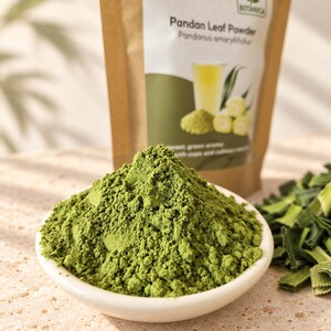 May include: A mound of vibrant green Pandan leaf powder sits in a small white bowl. Behind it is a brown paper pouch labeled Botanica Pandan Leaf Powder with an image of a glass of green liquid and fresh Pandan leaves. Chopped leaves are nearby.