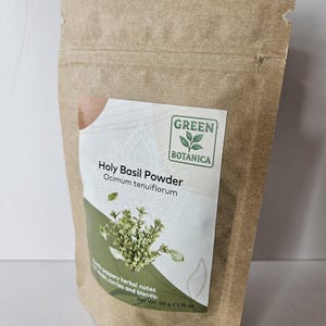 K&ouml;nnte beinhalten: Ein brauner Papiert&uuml;tchen mit Holy Basil Powder von Green Botanica. Das Etikett tr&auml;gt den Text "Holy Basil Powder" und "Ocimum tenuiflorum", mit einem Basilikumzweig und den Worten "pfeffrige Kr&auml;uternoten, Suppen, Currys und Mischungen." Das Nettogewicht betr&auml;gt 50 g.