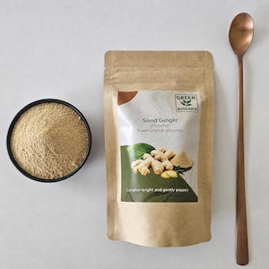 Op de afbeelding: Een bruin papieren zakje met gemalen zandgember van Green Botanica met het label Kaempferia galanga, een zwarte kom gevuld met het lichtbruine poeder en een lange koperkleurige metalen lepel op een neutraal oppervlak.