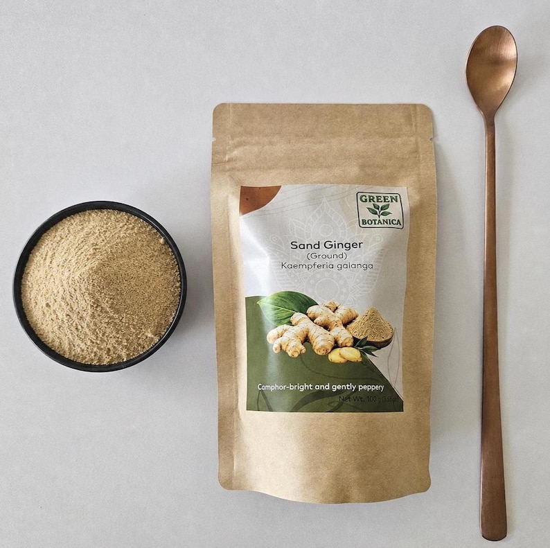 Op de afbeelding: Een bruin papieren zakje met gemalen zandgember van Green Botanica met het label Kaempferia galanga, een zwarte kom gevuld met het lichtbruine poeder en een lange koperkleurige metalen lepel op een neutraal oppervlak.