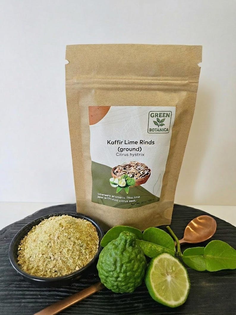 May include: A kraft paper bag of ground Kaffir lime rinds with the text "GREEN BOTANICA". A small bowl of ground lime rinds, a whole lime, a lime half, and a copper spoon are in the foreground. Green leaves and a whole lime are also visible.