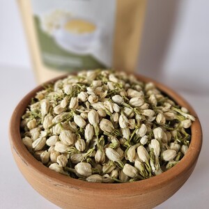 Peut inclure: Un bol en terre cuite rempli de fleurs de jasmin s&eacute;ch&eacute;es. Les fleurs sont blanches avec des tiges et des boutons verts. Un sac beige avec du texte est en arri&egrave;re-plan, flou. L'image est bien &eacute;clair&eacute;e et montre les d&eacute;tails des fleurs.