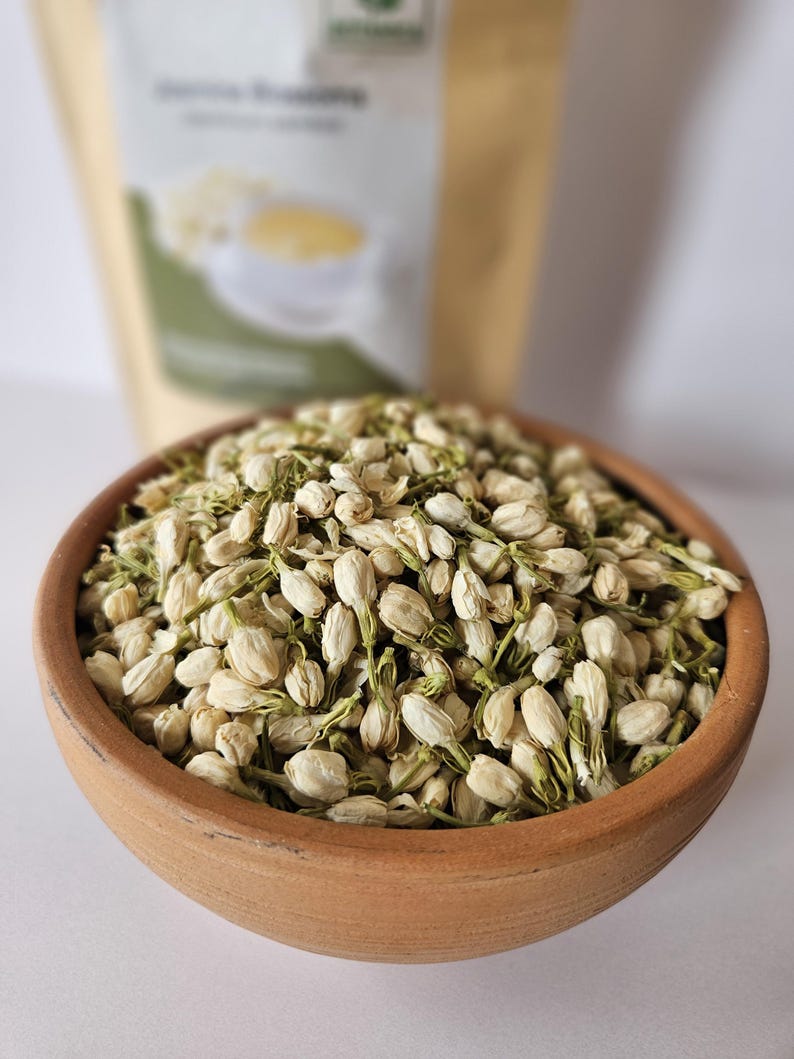 Peut inclure: Un bol en terre cuite rempli de fleurs de jasmin s&eacute;ch&eacute;es. Les fleurs sont blanches avec des tiges et des boutons verts. Un sac beige avec du texte est en arri&egrave;re-plan, flou. L'image est bien &eacute;clair&eacute;e et montre les d&eacute;tails des fleurs.