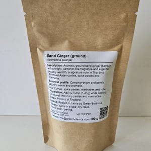 Op de afbeelding: Een bruine stazak van kraftpapier met 100 g gemalen zandgember. Het witte etiket bevat de tekst Sand Ginger (ground) Kaempferia galanga, een gedetailleerde productbeschrijving, gebruiksinstructies, herkomst en een QR-code.