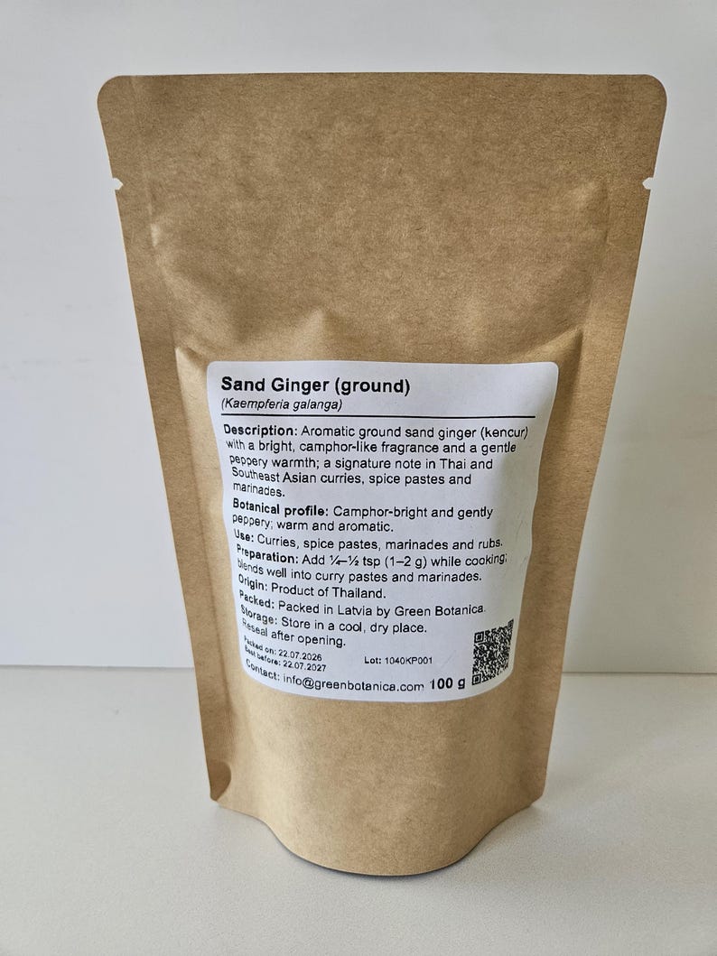 Op de afbeelding: Een bruine stazak van kraftpapier met 100 g gemalen zandgember. Het witte etiket bevat de tekst Sand Ginger (ground) Kaempferia galanga, een gedetailleerde productbeschrijving, gebruiksinstructies, herkomst en een QR-code.