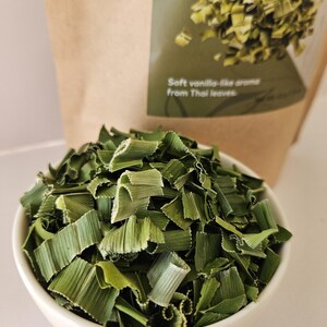 Może przedstawiać: Biała ceramiczna miska wypełniona suszonymi zielonymi liśćmi pandanu. Za miską brązowa papierowa torba z napisem "Soft vanilla-like aroma from Thai leaves" na zielonej etykiecie.