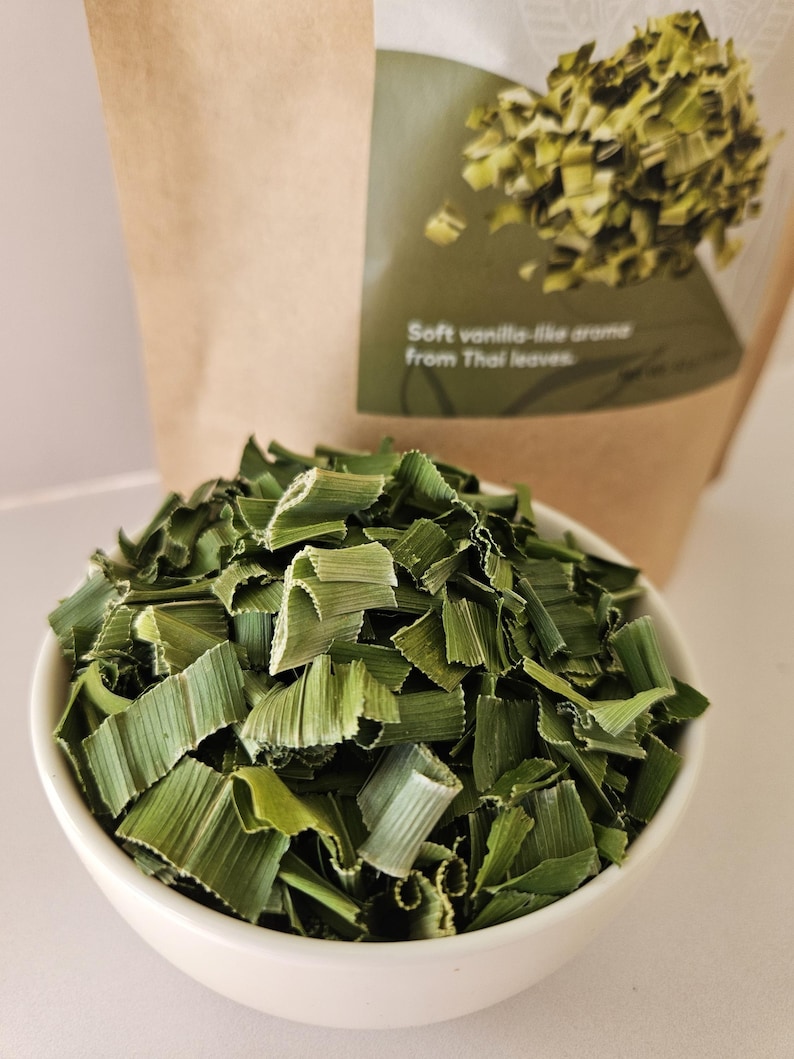 Może przedstawiać: Biała ceramiczna miska wypełniona suszonymi zielonymi liśćmi pandanu. Za miską brązowa papierowa torba z napisem "Soft vanilla-like aroma from Thai leaves" na zielonej etykiecie.